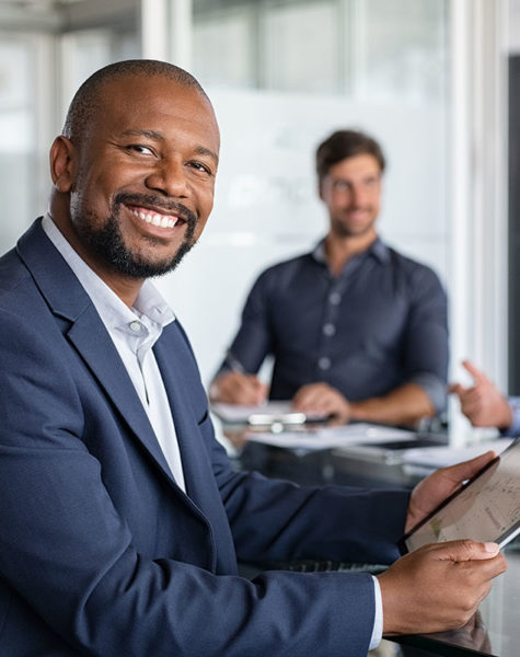 Proud mature black businessman smiling with colleagues sitting in a board room. Portrait of happy successful executive with team working in background while looking at camera during meeting. Success.