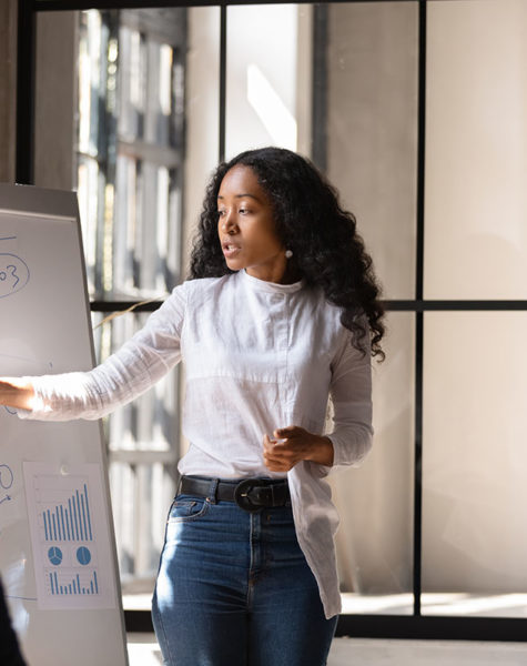 Young African American female coach or speaker make whiteboard presentation for diverse employees in office, focused biracial woman trainer present business project on flip chart at meeting